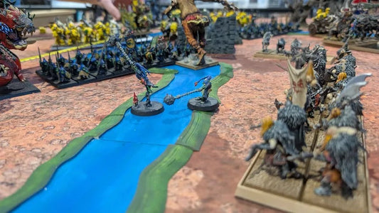 Painted fantasy armies clash across a tabletop battlefield, with two small figures standing in a bright blue river between them.